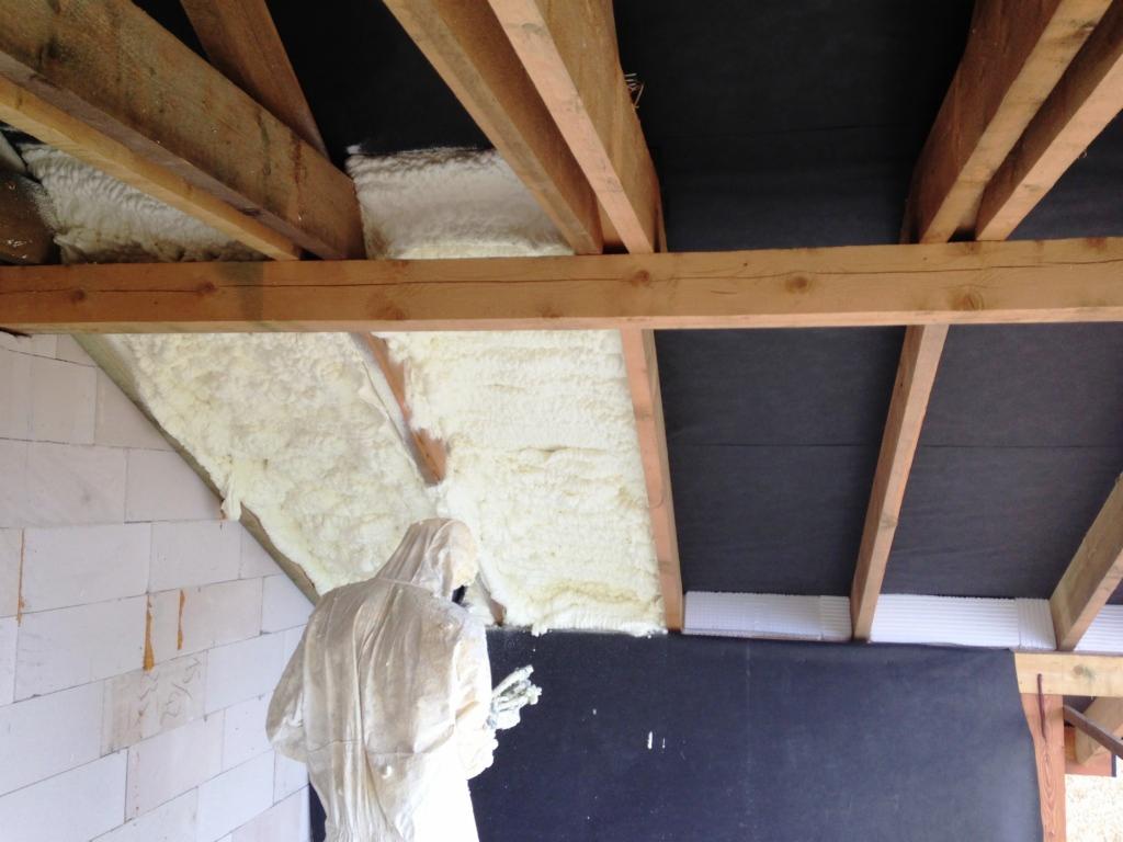 Prace izolacyjne poddasza: aplikacja jasnej piany izolacyjnej między krokwiami dachu, widoczna osoba w kombinezonie ochronnym podczas natrysku.