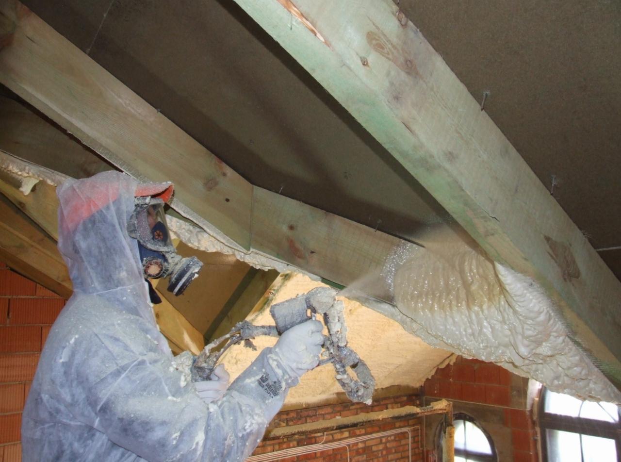 Pracownik w kombinezonie i masce ochronnej aplikuje pianę izolacyjną na drewnianą konstrukcję dachu, widoczne ceglane ściany i okno.