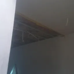 Niedokończona instalacja sufitu podwieszanego z widocznymi przewodami i metalową konstrukcją, przy ścianie z widocznymi śladami po taśmie malarskiej.