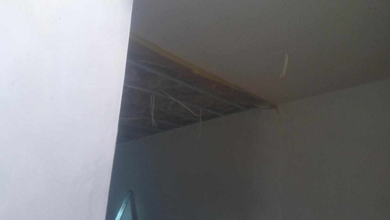 Niedokończona instalacja sufitu podwieszanego z widocznymi przewodami i metalową konstrukcją, przy ścianie z widocznymi śladami po taśmie malarskiej.