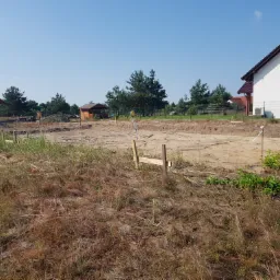 Wytyczanie fundamentów pod budowę domu jednorodzinnego, widoczny niwelator na statywie, drewniane paliki i wykopane fundamenty, w tle zabudowa mieszkalna.