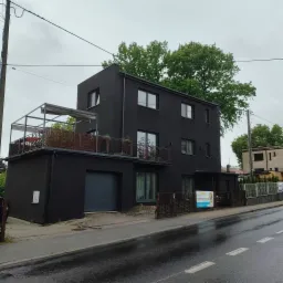 Nowoczesny, dwupiętrowy dom w czarnym kolorze z balkonem na pierwszym piętrze, widok z ulicy w pochmurny dzień. Minimalistyczna bryła budynku.