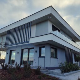 SALAMON ELEWACJE OCIEPLANIE - Nowoczesny, dwukondygnacyjny dom z płaskim dachem i szarymi panelami elewacyjnymi. Widoczne pionowe, ciemne lamele, szklana balustrada balkonu i starannie zagospodarowany ogród.