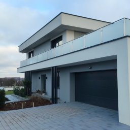 SALAMON ELEWACJE OCIEPLANIE - Nowoczesny dom z płaskim dachem i balkonem z szklaną balustradą. Elewacja w odcieniach szarości, brama garażowa segmentowa. Kostka brukowa na podjeździe.