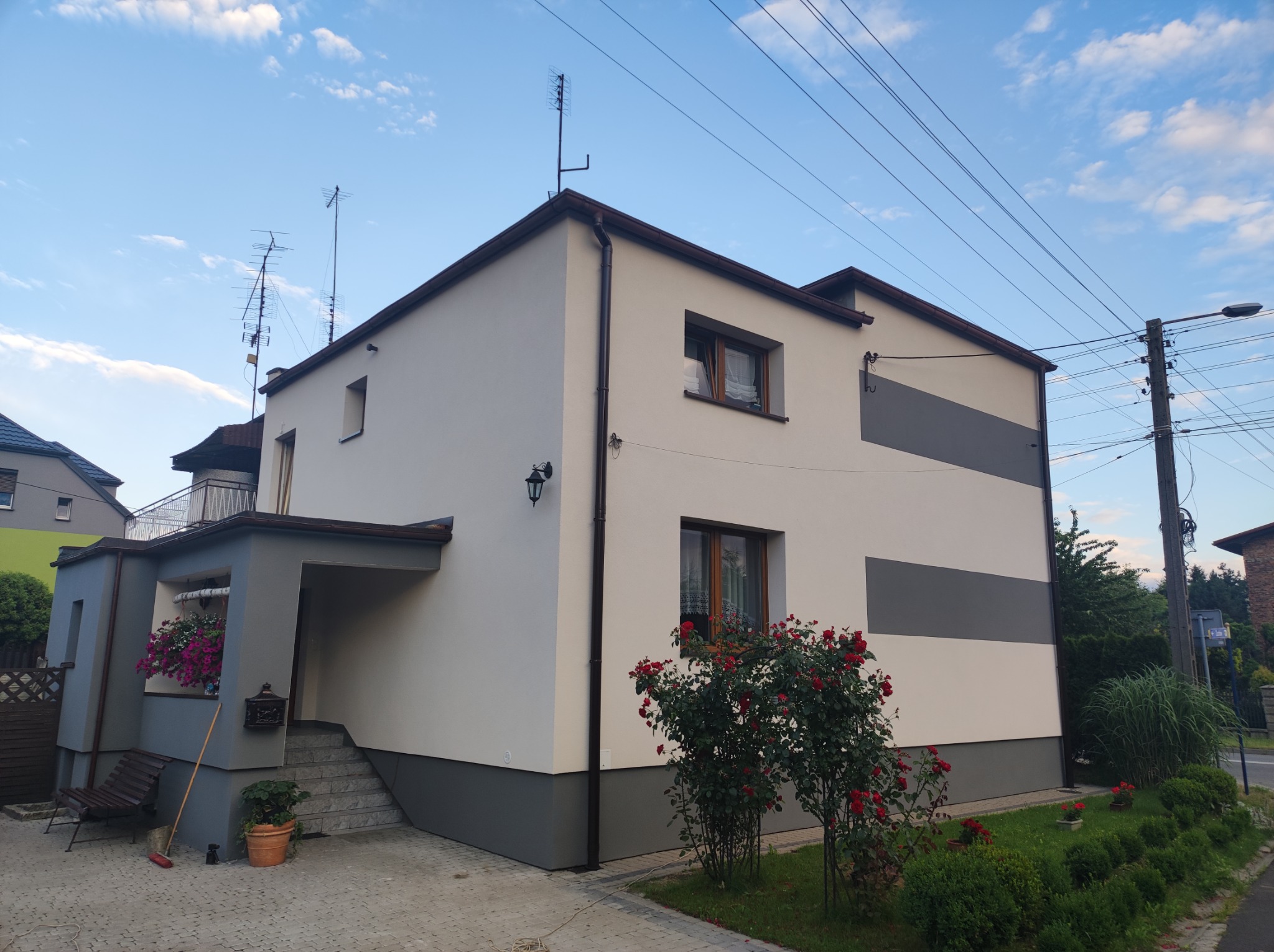 Dwukondygnacyjny dom jednorodzinny z jasną elewacją, szarymi akcentami i brązowymi ramami okiennymi, otoczony zielenią i kwiatami, sfotografowany w słoneczny dzień.