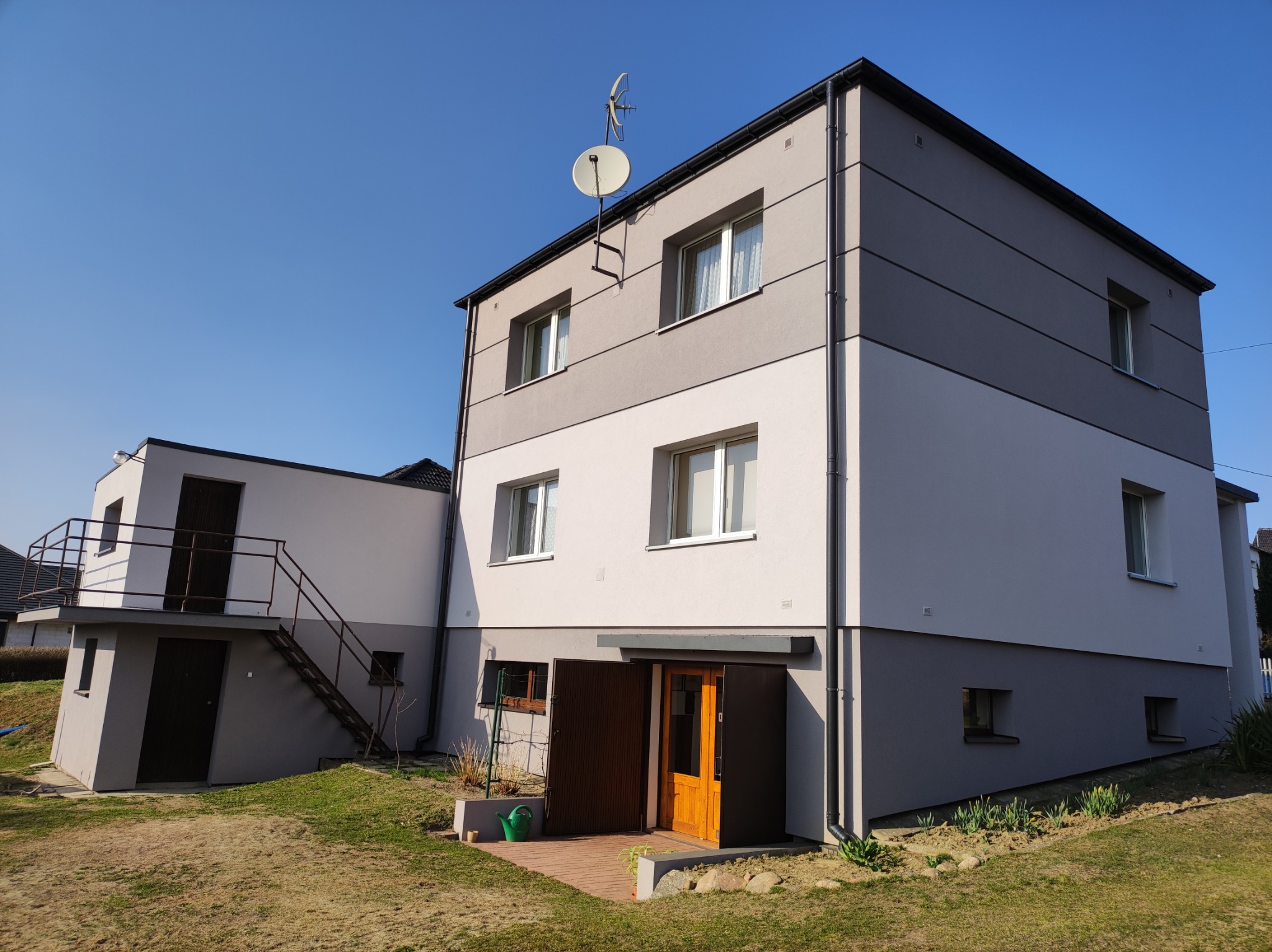 Dwukondygnacyjny dom z elewacją w odcieniach szarości i bieli, z anteną satelitarną na dachu, dodatkowy budynek z zewnętrznymi schodami, widoczne kwiaty i konewka przy wejściu.