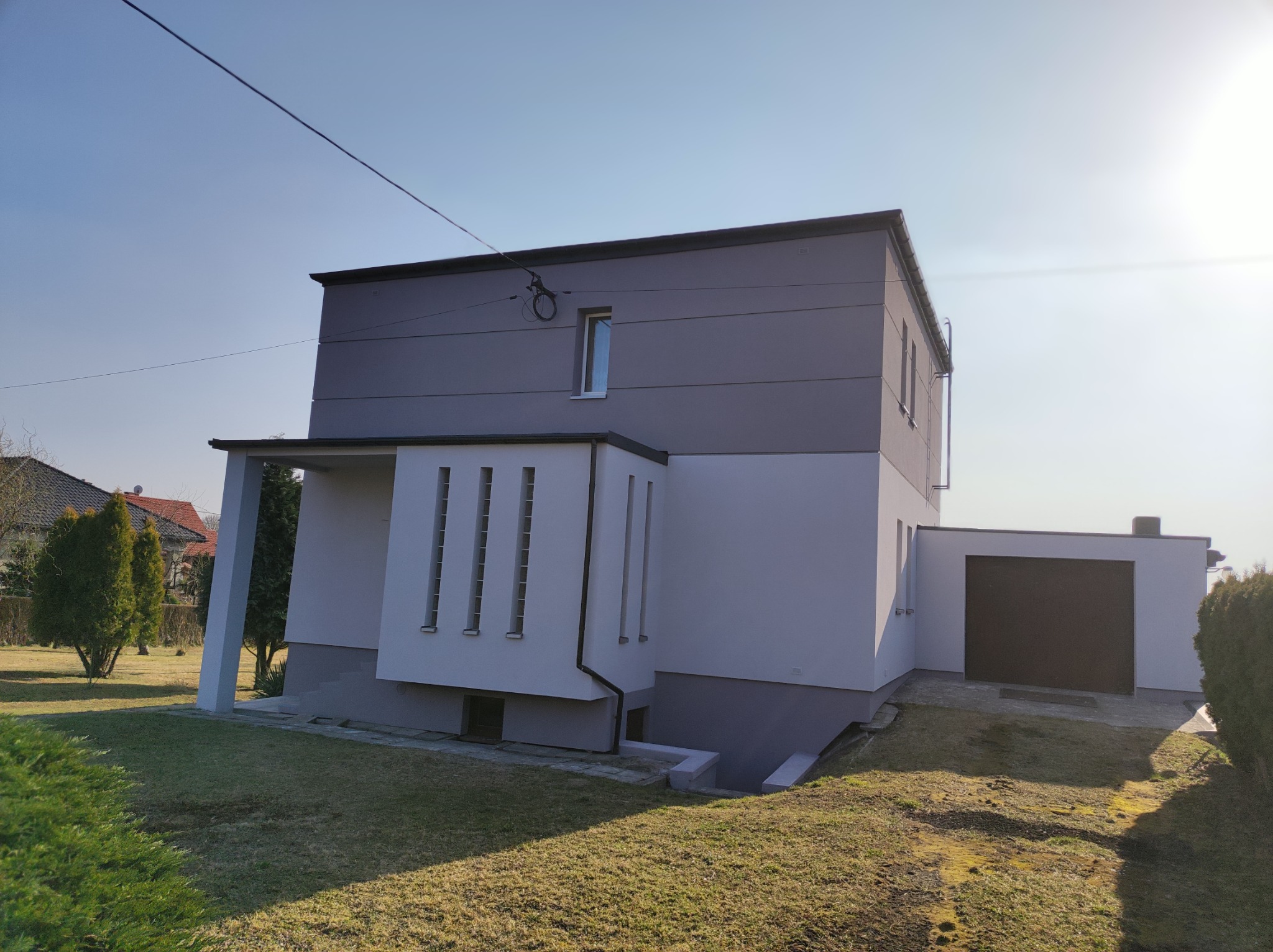 Dwukondygnacyjny dom z płaskim dachem, pomalowany w odcieniach szarości i bieli, z garażem dobudowanym z boku, widoczny z perspektywy trawnika w słoneczny dzień.
