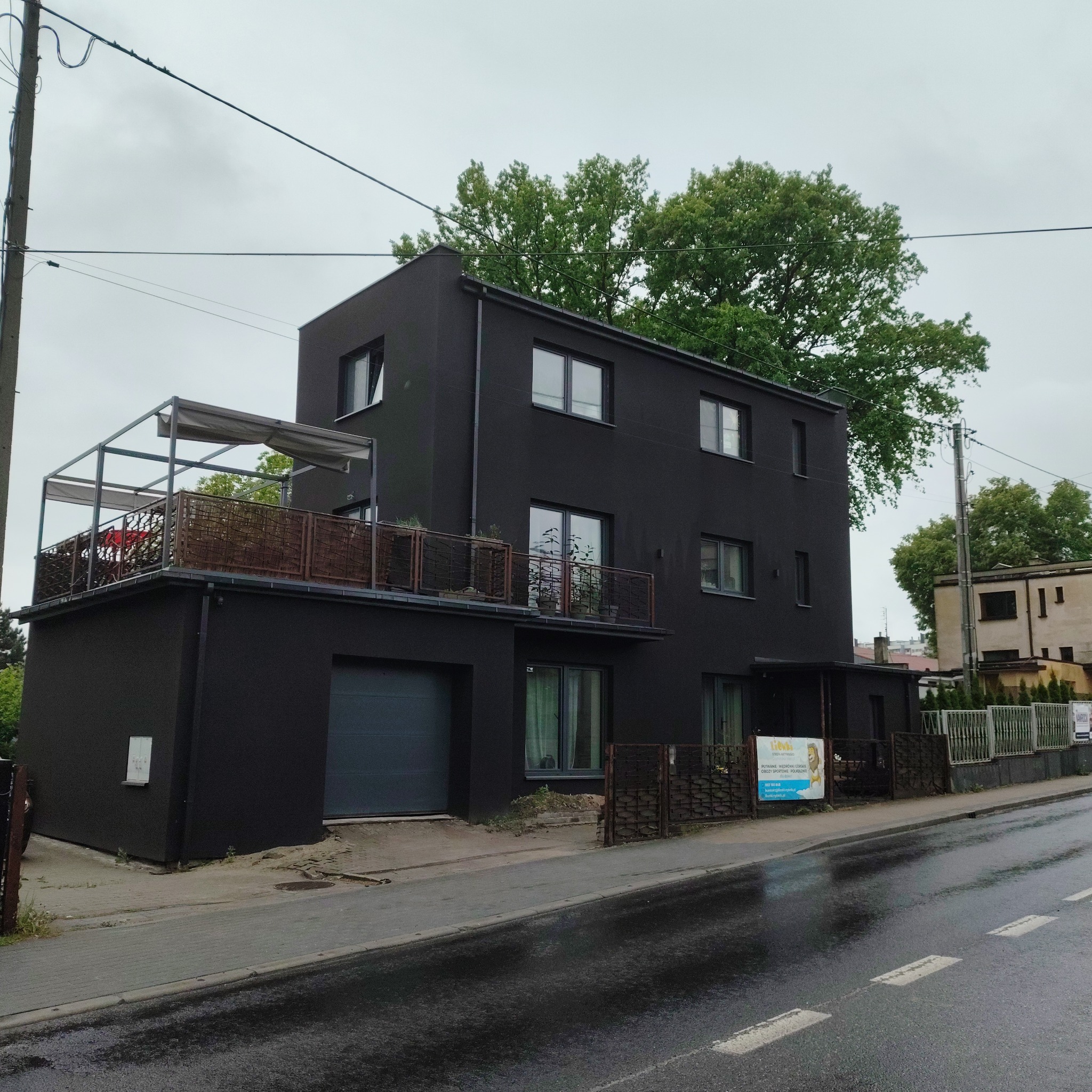 Nowoczesny, dwupiętrowy dom w czarnym kolorze z balkonem na pierwszym piętrze, widok z ulicy w pochmurny dzień. Minimalistyczna bryła budynku.
