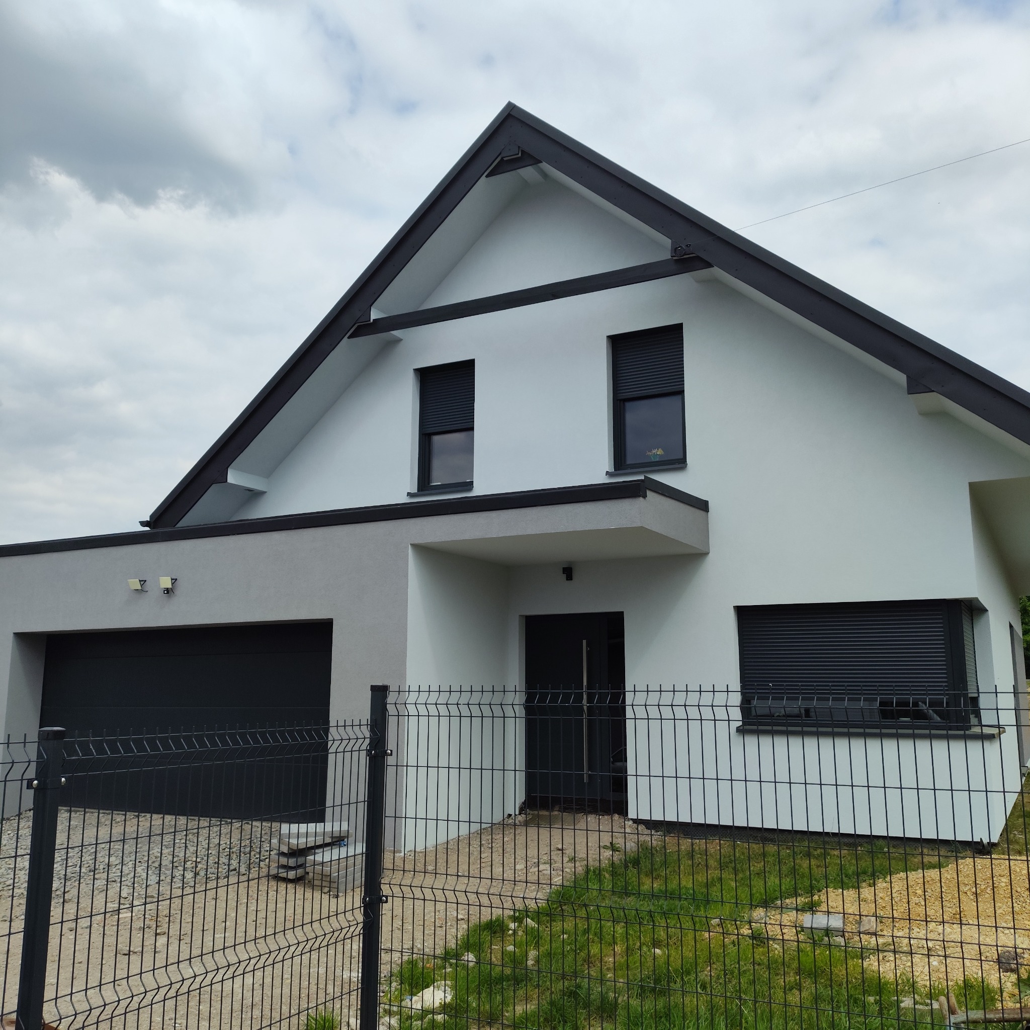 Nowoczesny dom z białą elewacją, czarnymi akcentami i szarą częścią garażową, ogrodzony metalową siatką. Widoczne okna z roletami.
