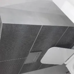 Szare płytki z dekoracyjnym wzorem na ścianie w łazience, widoczny fragment toalety z deską i przycisk spłuczki.