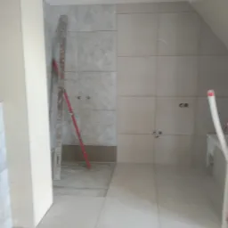 Łazienka w trakcie remontu z ułożonymi płytkami podłogowymi i ściennymi, widoczna poziomica oparta o ścianę i przygotowane otwory na armaturę.