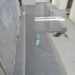 Układanie szarych płytek podłogowych z widocznymi krzyżykami dystansowymi, butelka wody na płytkach, w tle widoczne pomieszczenie w trakcie remontu.