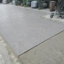 Nowa, szara posadzka przemysłowa z antypoślizgową fakturą, częściowo pokryta świeżym jastrychem, w tle widoczne maszyny i wózki transportowe.