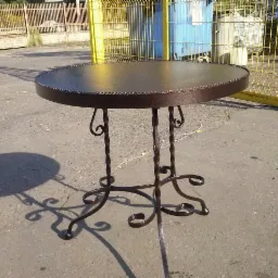 Okrągły metalowy stolik z dekoracyjnymi, spiralnymi nogami, stojący na betonowym podłożu w plenerze, z widocznym cieniem i metalowym ogrodzeniem w tle.