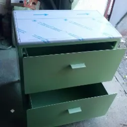 Zielona metalowa szafka z dwiema szufladami, g&oacute;rna szuflada wysunięta, na g&oacute;rze szafki arkusz blachy z folią ochronną i strzałkami, widoczne otoczenie warsztatowe.