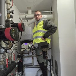 PU MARCO - Instalacje gazowe - Instalator w kamizelce, z miernikiem, kontroluje instalację gazową w kotłowni. Widoczne rury, zawory, pompa obiegowa i piec gazowy. Wąskie pomieszczenie.