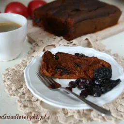 Kawa w białym kubku obok kromki ciemnego ciasta z suszonymi śliwkami i żurawiną na białym talerzyku, całość na beżowej serwetce z napisem www.poradniadietetyki.pl.