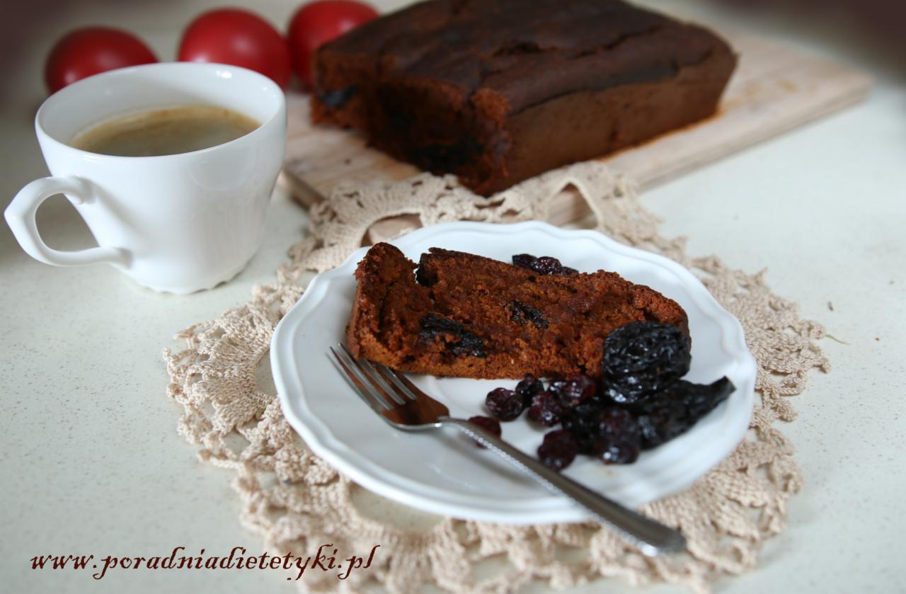 Kawa w białym kubku obok kromki ciemnego ciasta z suszonymi śliwkami i żurawiną na białym talerzyku, całość na beżowej serwetce z napisem www.poradniadietetyki.pl.