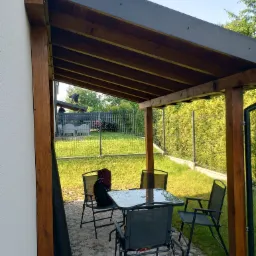 Drewniana altana z metalowym dachem, wyposażona w szary stół i krzesła na kostce brukowej, z widokiem na zielony ogród z siatką ogrodzeniową w tle.