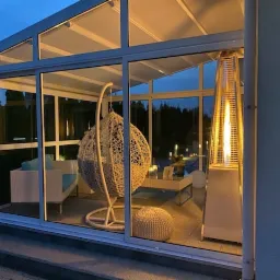 Zadaszony taras z białymi ramami, widoczne rattanowe meble ogrodowe, wiszące krzesło i stojący grzejnik tarasowy z płomieniem.