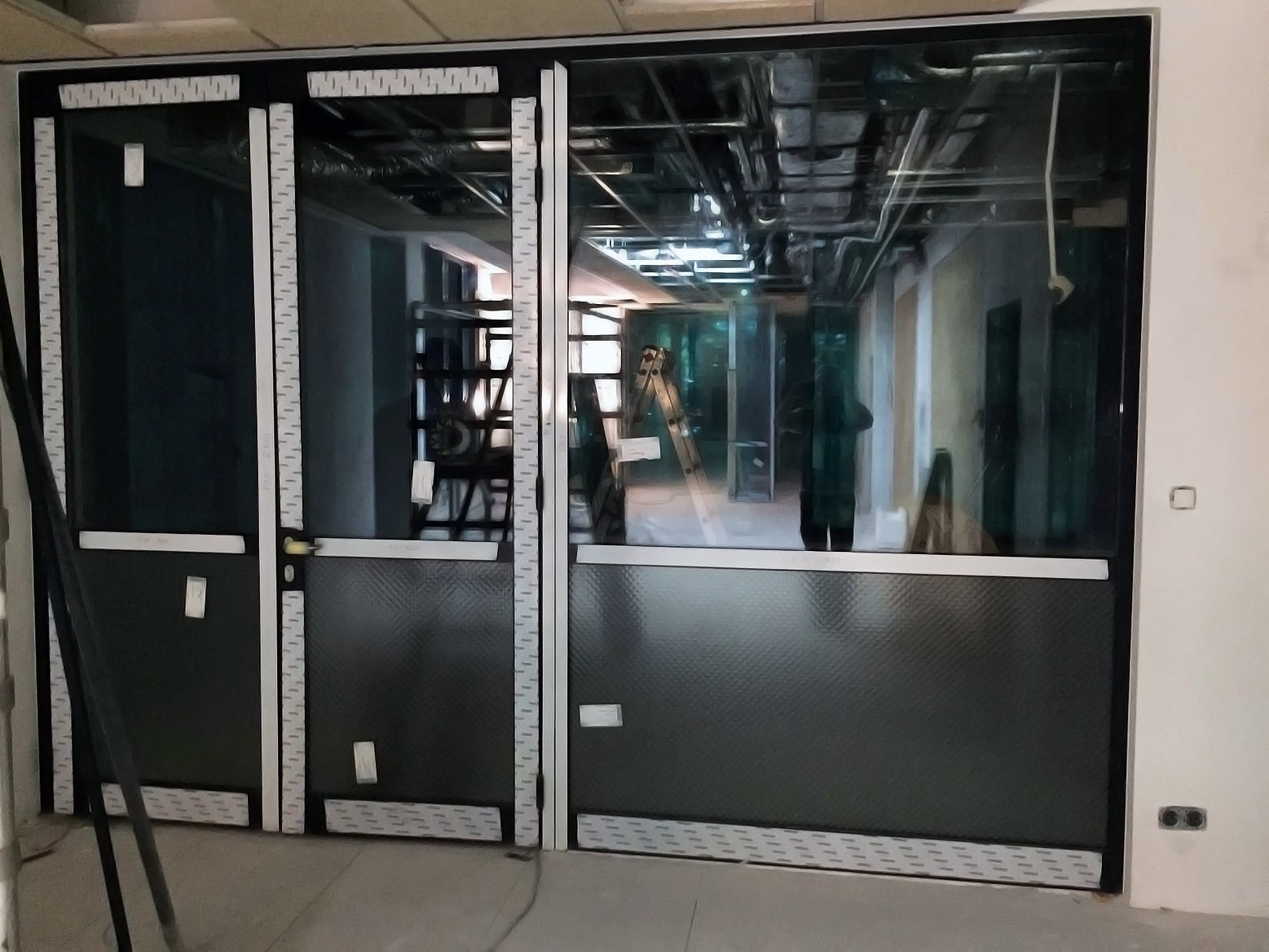 Wewnętrzna zabudowa aluminiowa z czarnym panelem dolnym i szkłem, widoczne instalacje pod sufitem i drabina w tle, zabezpieczona taśmą ochronną.