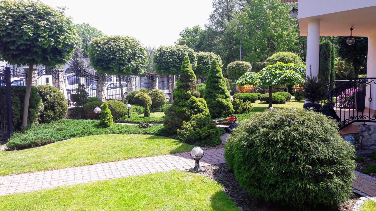 Starannie utrzymany ogród z różnorodnymi, formowanymi drzewami i krzewami, oświetlony lampami ogrodowymi wzdłuż ścieżki z kostki brukowej. W tle widoczny dom z kolumnami i ozdobną balustradą.