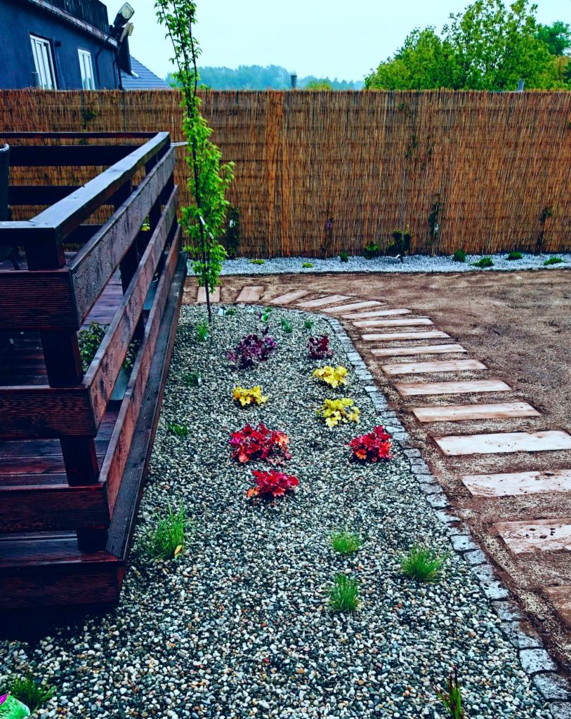 Ogród z drewnianym tarasem, ścieżką z płyt i rabatą wysypaną żwirem z nasadzeniami kolorowych kwiatów oraz młodego drzewka, otoczony bambusowym płotem.