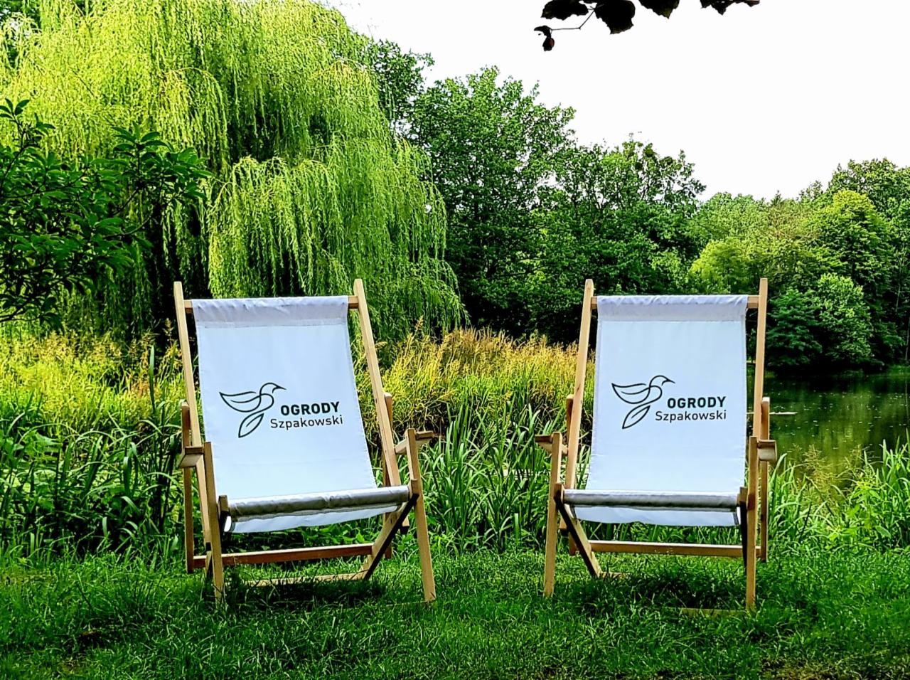 Dwa drewniane leżaki z logo firmy ogrodniczej na tle zielonego ogrodu z jeziorkiem w tle.