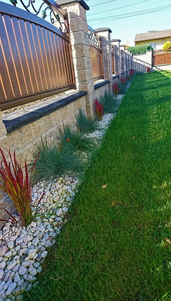 Ogród z nowoczesnym ogrodzeniem z brązowymi panelami i kamiennymi słupkami, obsadzony ozdobnymi trawami i białymi kamykami, przylegający do zadbanego trawnika.