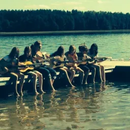 Grupa dzieci siedzi na pomoście nad jeziorem, grając na gitarach akustycznych, z nogami zanurzonymi w wodzie, w tle las i błękitne niebo.