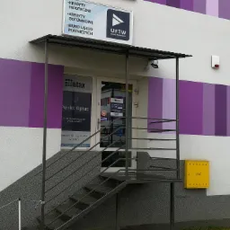 Wejście do biura UFTW Usługi Finansowe z widoczną tablicą informacyjną o kredytach hipotecznych i gotówkowych, schody z metalową barierką i żółtą skrzynką gazową na ścianie budynku...