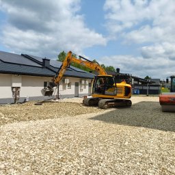 SIGMA Łukasz Zątek - Żółta koparka JCB na placu budowy, przygotowuje podłoże z białego kruszywa. W tle budynek z panelami słonecznymi i walec HAMM.