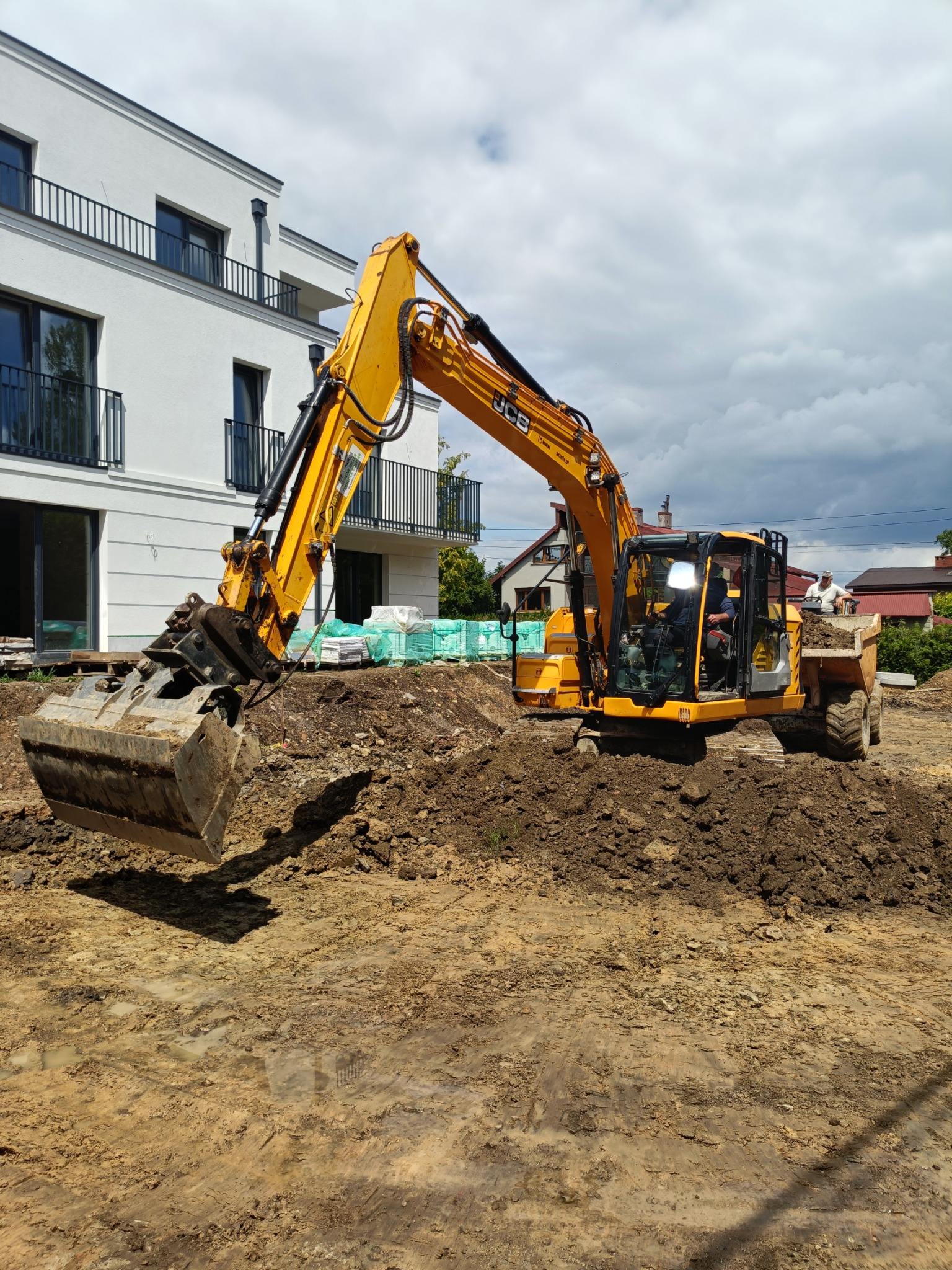 Żółta koparka JCB na placu budowy przed nowoczesnym, białym budynkiem z balkonami. W tle widoczne materiały budowlane i pochmurne niebo.