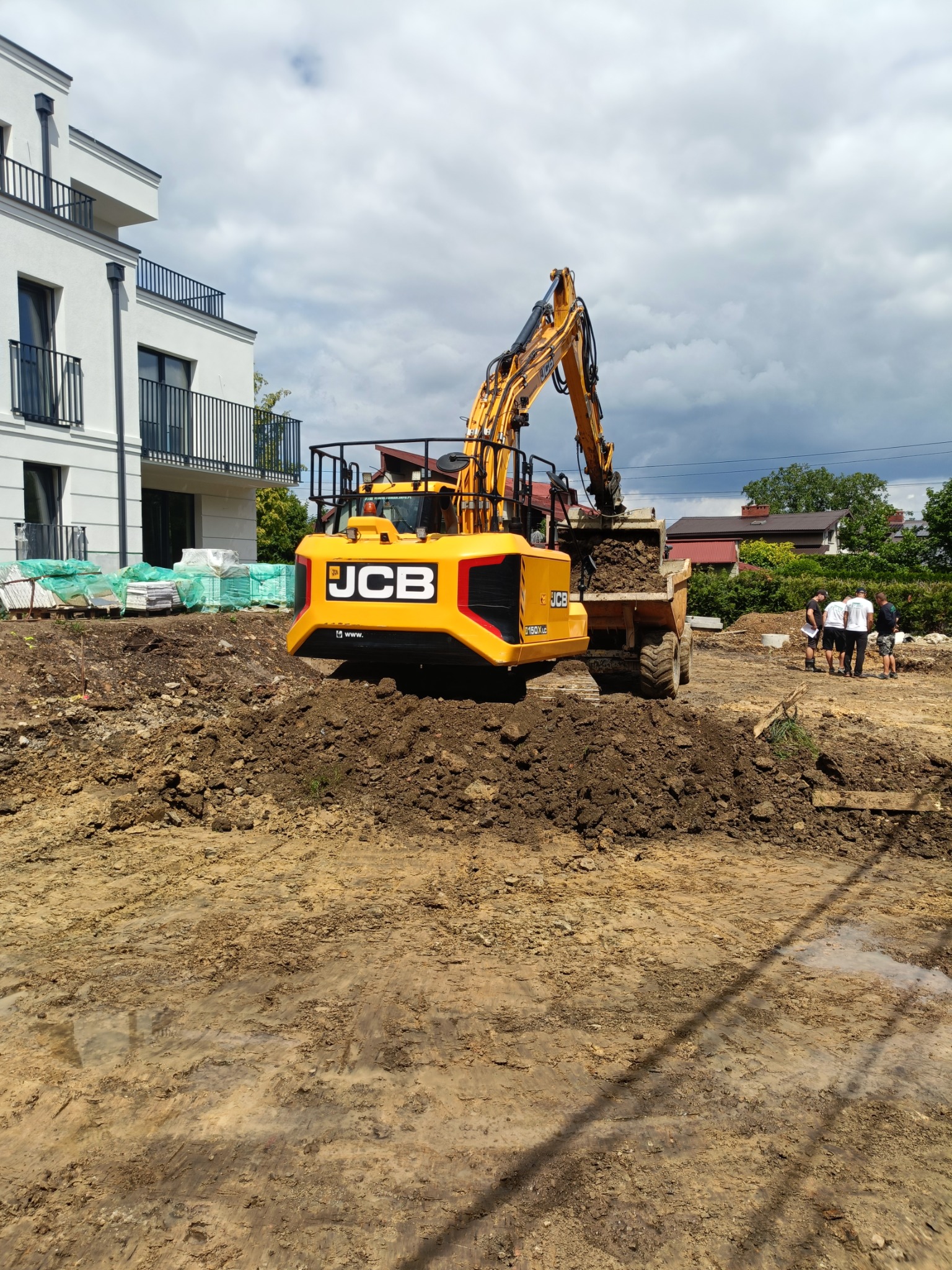 Żółta koparka JCB ładuje ziemię do wywrotki na placu budowy, w tle biały budynek z balkonami i grupa osób.