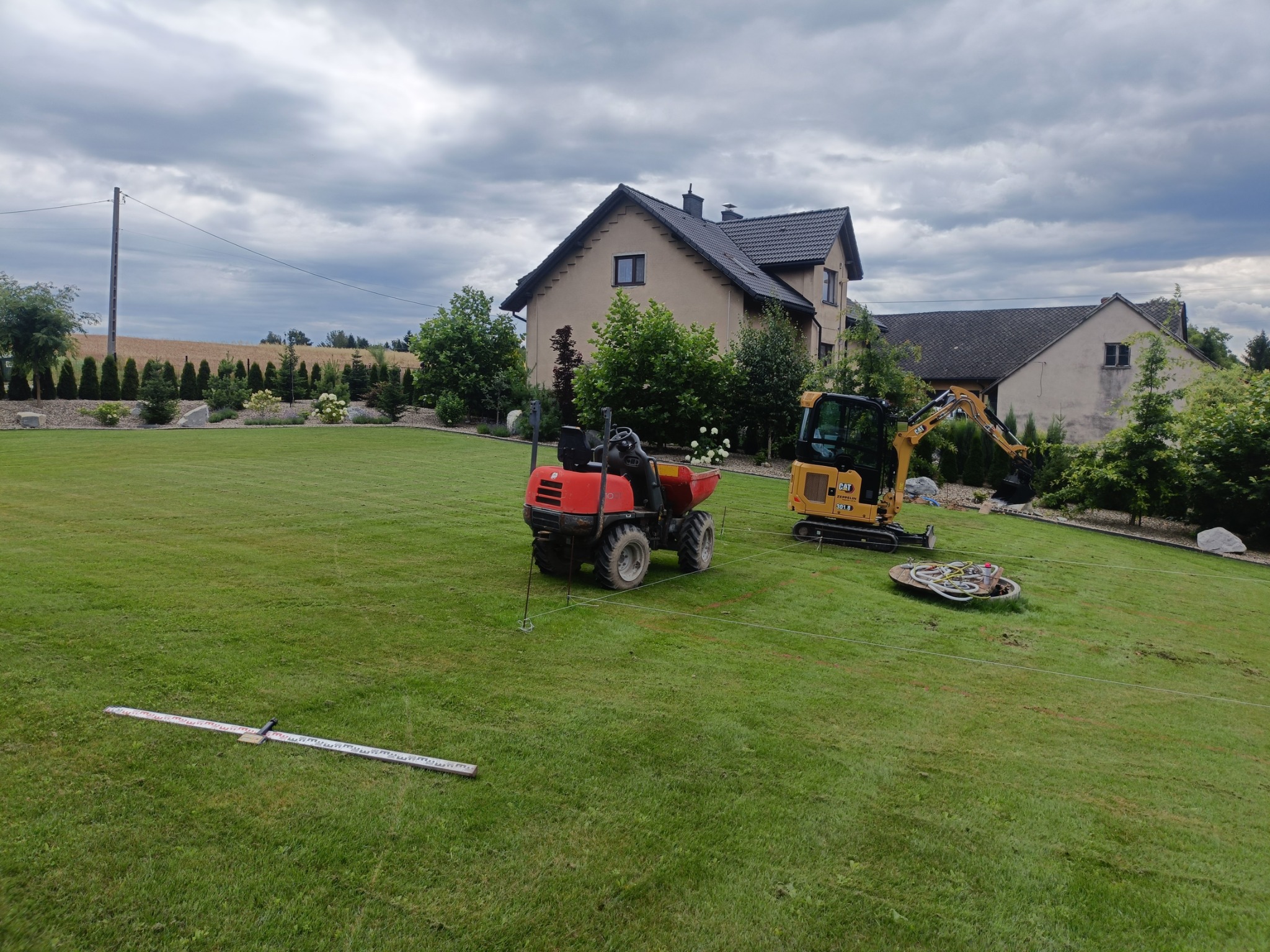 Przygotowanie terenu: mini wywrotka i koparka na trawniku, wytyczone sznurki, łata niwelacyjna, dom w tle. Prace ziemne przed założeniem ogrodu.
