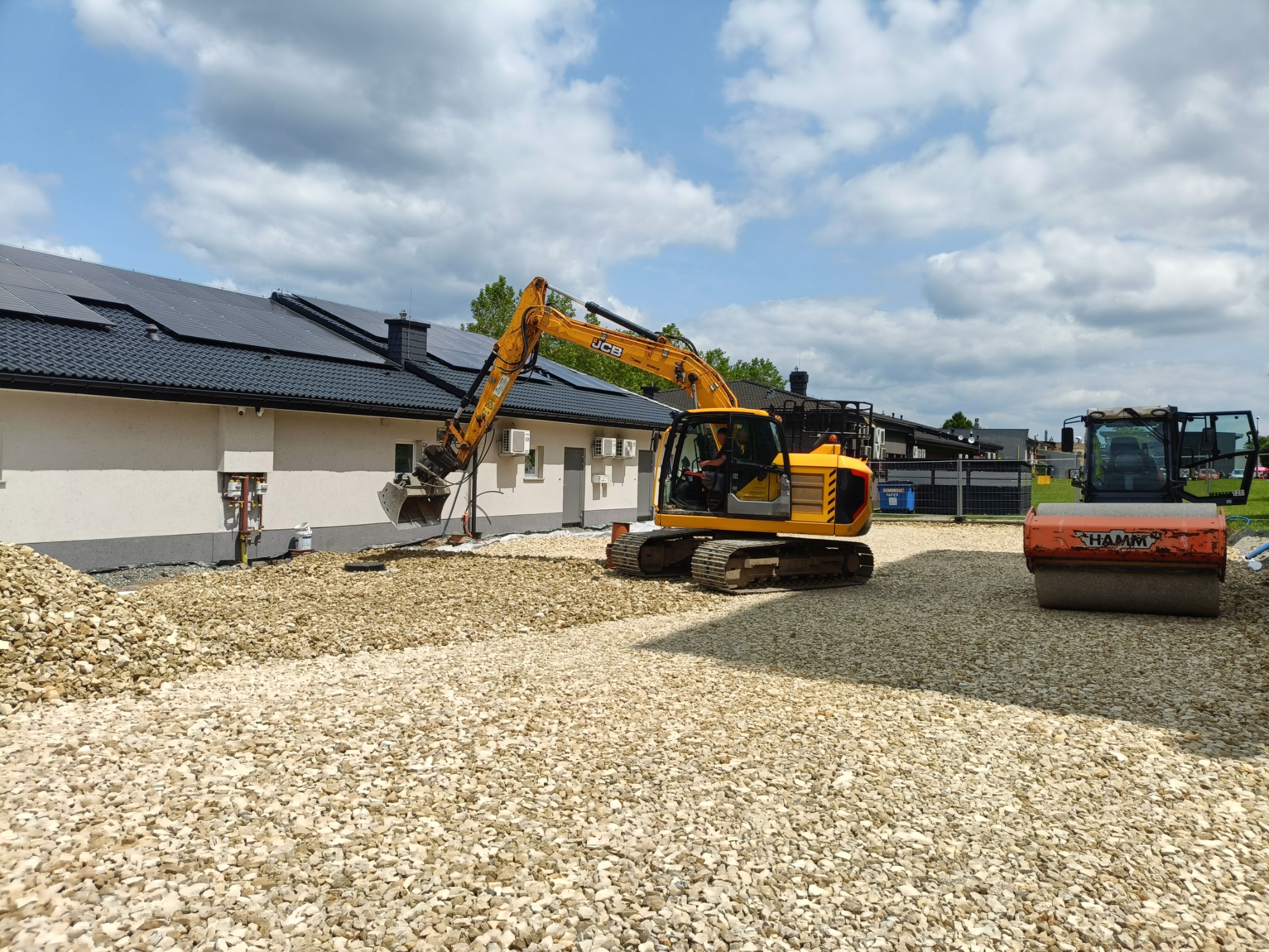 Żółta koparka JCB na placu budowy, przygotowuje podłoże z białego kruszywa. W tle budynek z panelami słonecznymi i walec HAMM.