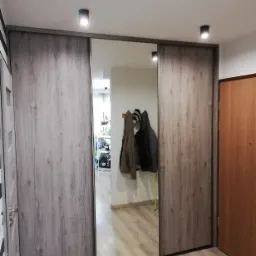 Szafa wnękowa z przesuwnymi drzwiami z lustrem, imitacja drewna, oświetlenie sufitowe punktowe, widoczne ubrania w odbiciu lustrzanym.