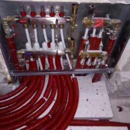 Rozdzielacz ogrzewania podłogowego z czerwonymi rurami w trakcie montażu, widoczne zawory, manometry i białe rurki doprowadzające w osłonie.