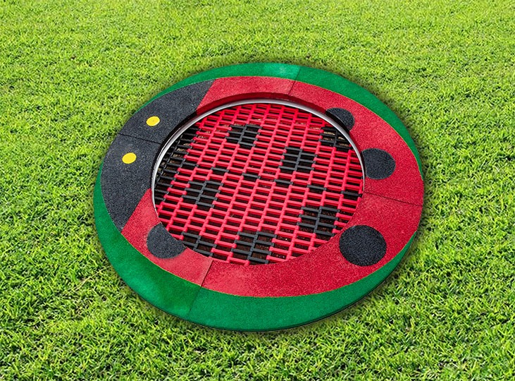 Trampolina ziemna w kształcie biedronki z czerwono-czarną siatką na zielonym trawniku.