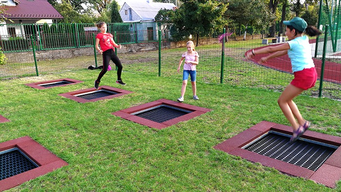 Dzieci skaczą na wbudowanych trampolinach na trawiastym terenie, otoczonych zielonym ogrodzeniem i budynkami w tle.
