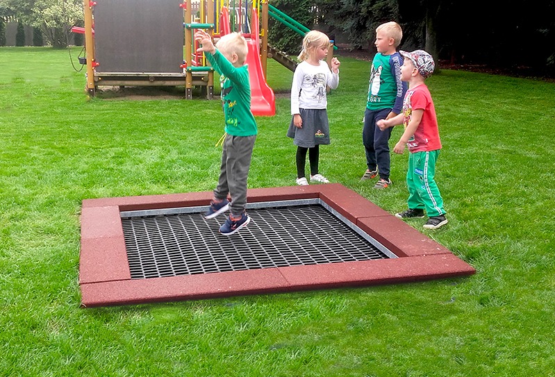 Chłopiec w zielonej koszulce skacze na wbudowanej w ziemię, kwadratowej trampolinie z metalową siatką, otoczonej czerwoną gumową obudową, na tle zielonego trawnika i innych dzieci.