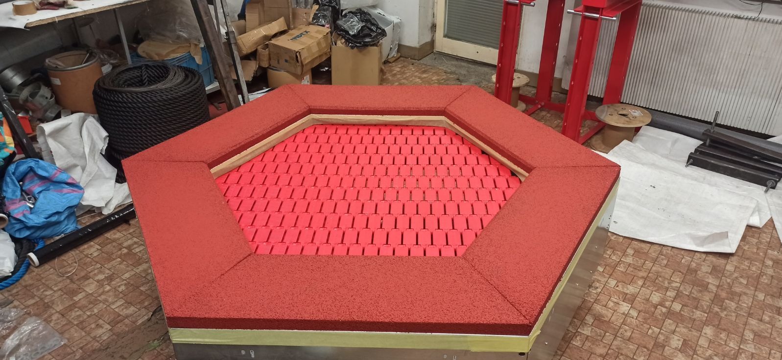 Sześciokątna konstrukcja z czerwonej gumy z wypełnieniem z czerwonych elementów w kształcie litery T, prawdopodobnie element wyposażenia placu zabaw, w trakcie montażu w warsztacie.