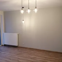 Pomalowane na szaro wnętrze pokoju z panelami podłogowymi, białym grzejnikiem i nowoczesną lampą wiszącą z czterema szklanymi kloszami.
