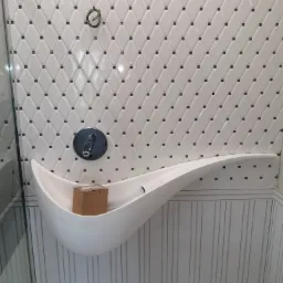 Biała, nowoczesna półka w kształcie fali zamontowana w kabinie prysznicowej z biało-czarną mozaiką diamentową na ścianach, widoczny kran i wystające przewody elektryczne.