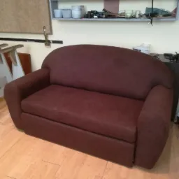 Brązowa sofa dwuosobowa na drewnianej podłodze, z widocznymi narzędziami i materiałami na półce w tle, sugerującymi warsztat.