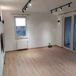 Pusty pokój z jasnymi panelami podłogowymi, białymi ścianami, grzejnikiem pod oknem, oświetleniem szynowym na suficie i lampami ściennymi.