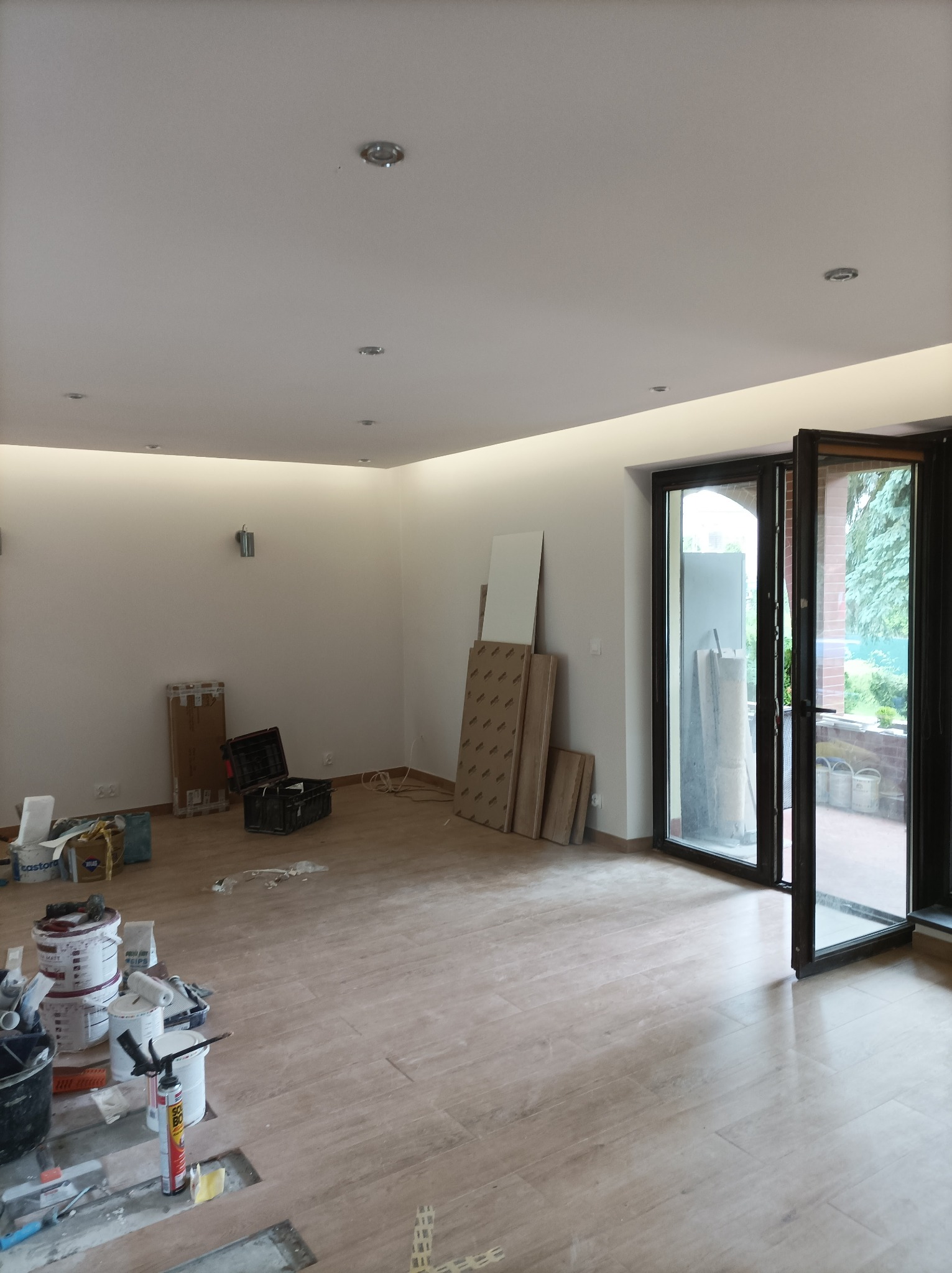 Wnętrze w trakcie prac wykończeniowych: widoczne narzędzia, puszki z farbą, płyty gipsowe oparte o ścianę, częściowo położona podłoga z płytek imitujących drewno, otwarte drzwi balkonowe.
