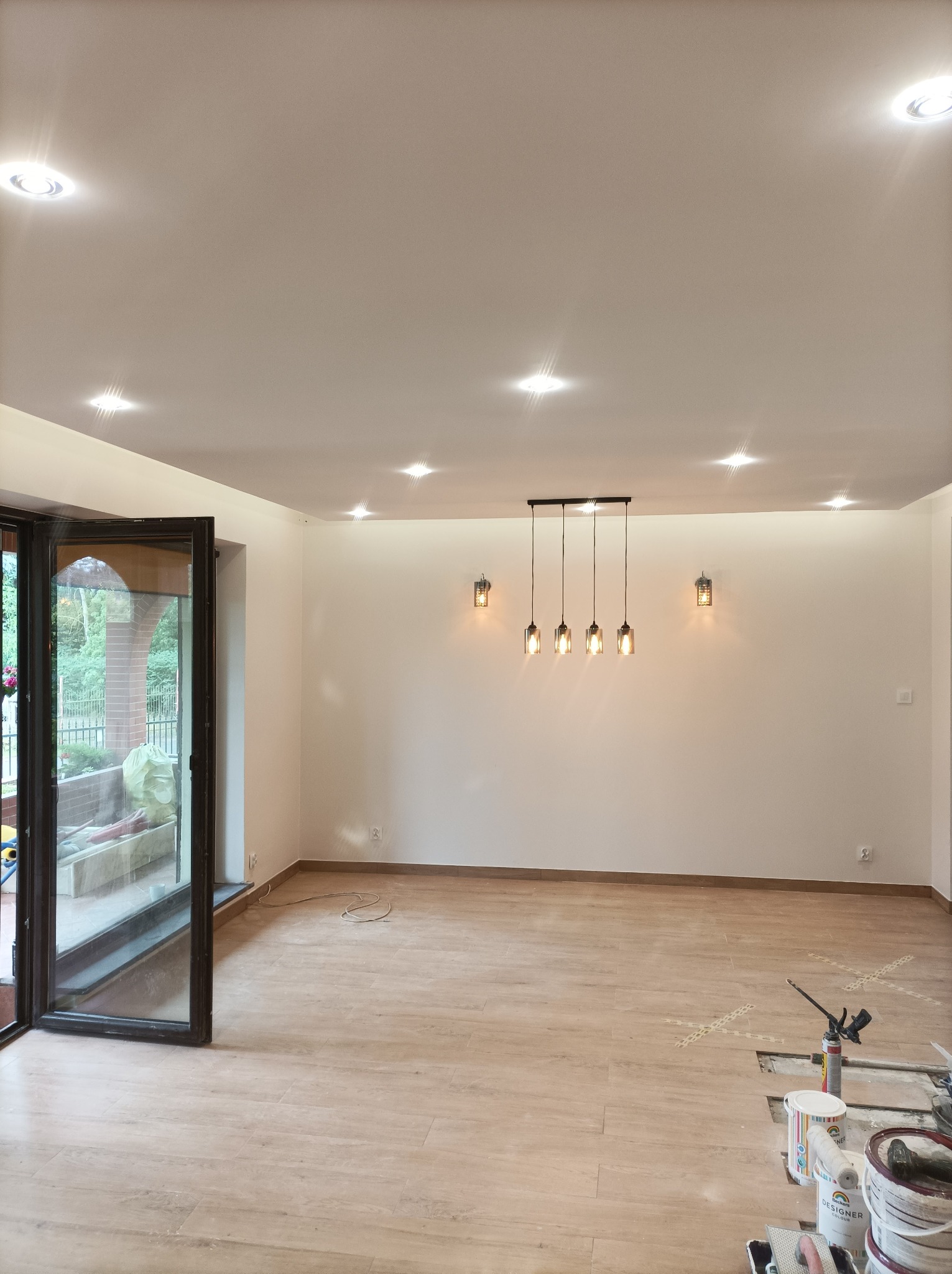 Jasne wnętrze w trakcie prac wykończeniowych: panele podłogowe ułożone, ściany pomalowane na biało, widoczne puszki z farbą i narzędzia malarskie w rogu pomieszczenia, wiszące lampy w stylu...