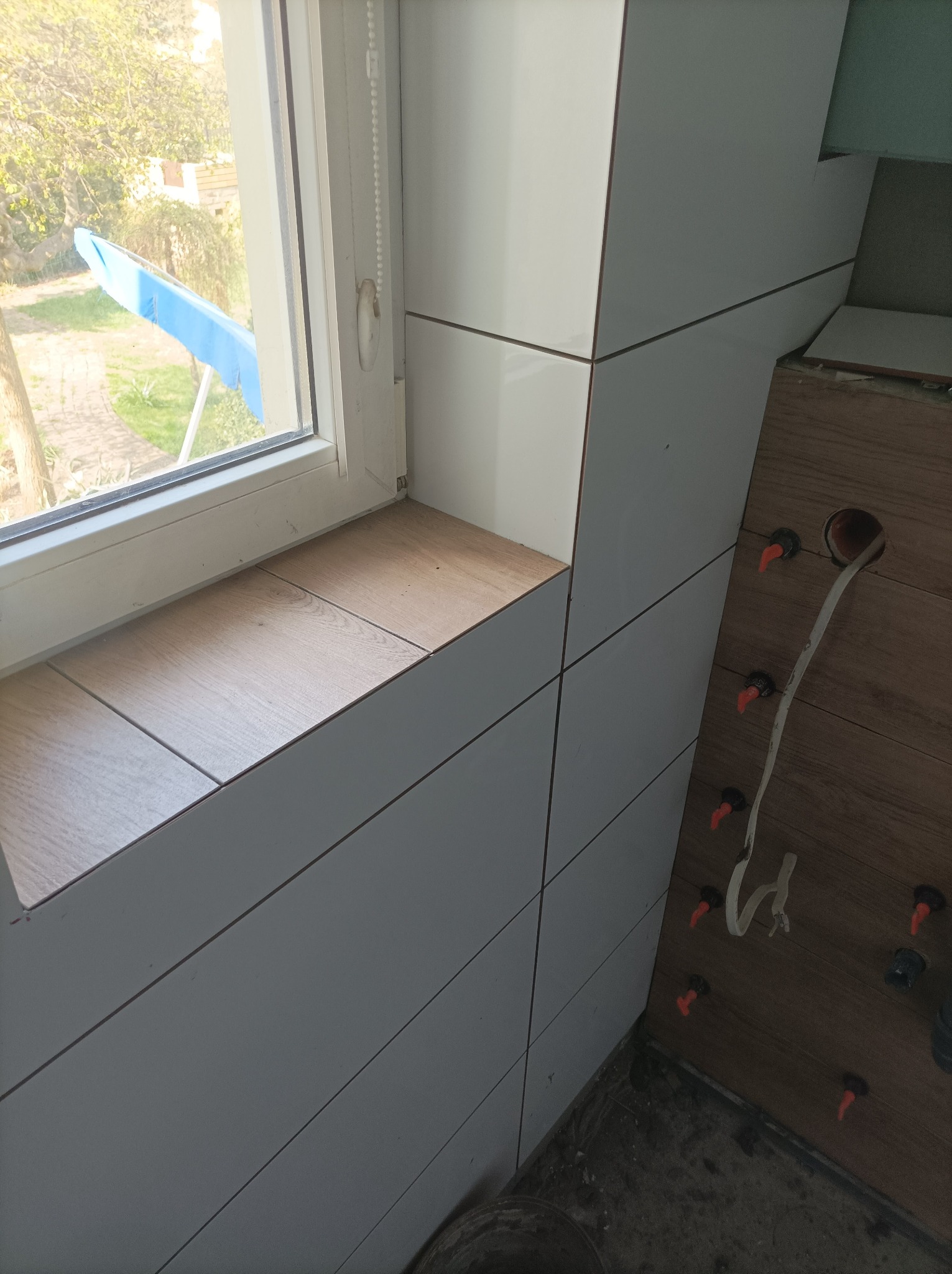 Białe i drewnopodobne płytki ułożone na ścianie i parapecie okiennym, widoczne narzędzia glazurnicze i przewód elektryczny.