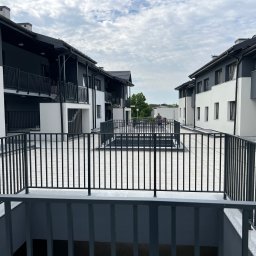 ITB Biuro Inżynierskie Piotr Kurasiński - Nowoczesne osiedle budynków wielorodzinnych z balkonami i wewnętrznym dziedzińcem wyłożonym kostką, widok z wyższego poziomu na metalowe balustrady i osobę siedzącą w oddali.